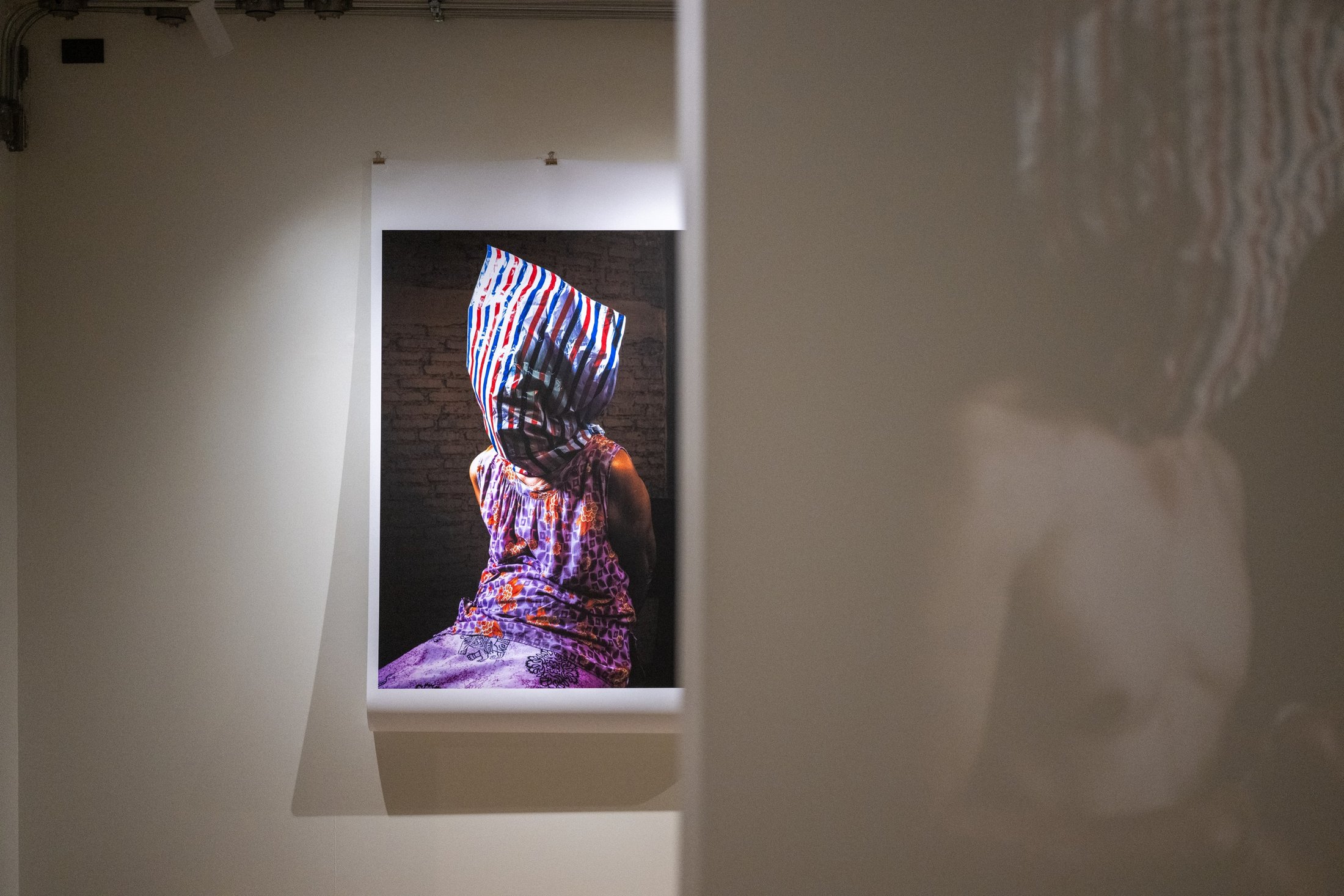 Installation view of Delusional: a large photographic print of a figure with a striped plastic bag over their head, hung on a white wall